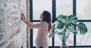 Woman adjusting a wall-mounted thermostat next to a large window and a potted plant