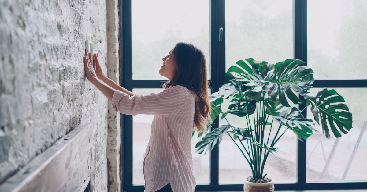Woman adjusting a wall-mounted thermostat next to a large window and a potted plant